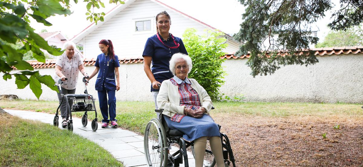 Bilden visar två skötare som hjälper två boende på promenad utomhus, en rullstolsburen och en med rullator.