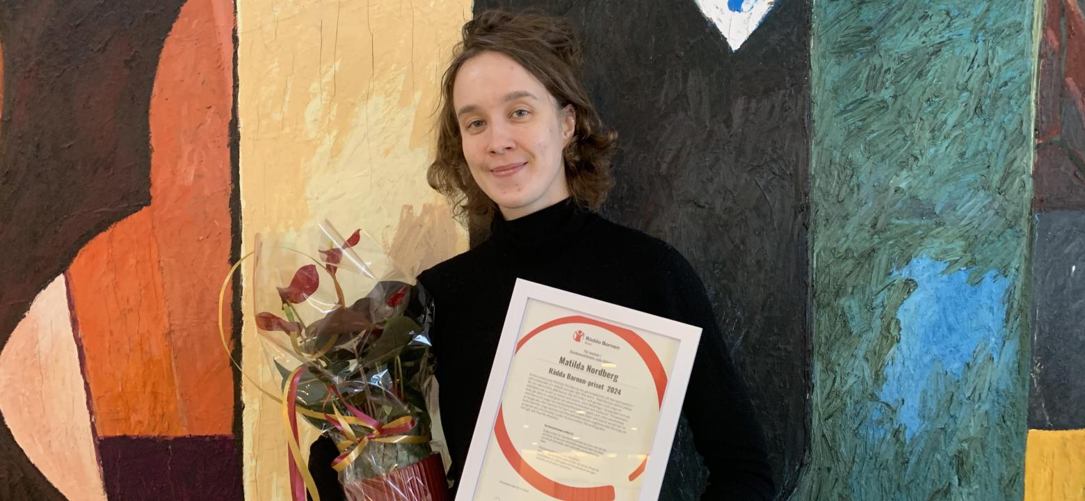 Foto på kulturproducent Matilda Nordberg, framför stadsbibliotekets konstverk av Johan Scott "Samtal och utan titel" Hon håller i sitt diplom och en röd blomma. 